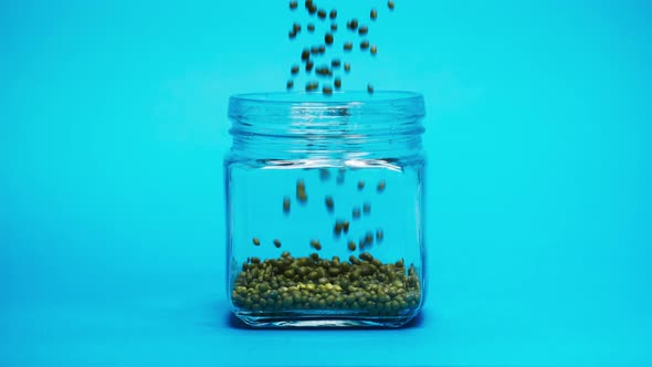 Closeup of Falling Down Green Mung Beans Into Glass Jar on Blue Background