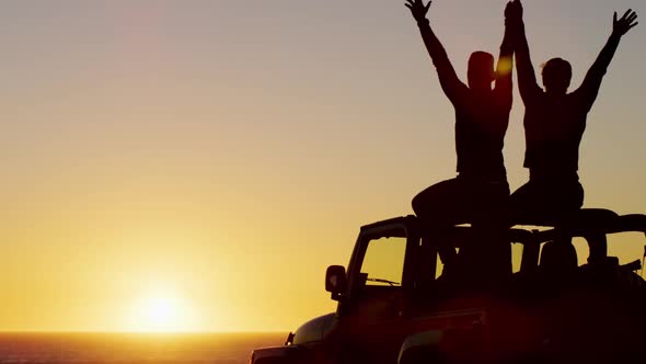 Happy caucasian gay male couple sitting on car raising arms and holding hands at sunset on the beach