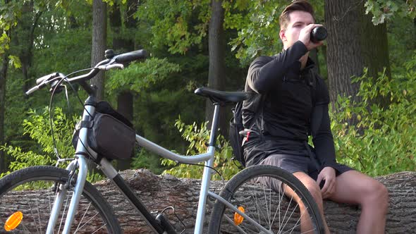 A Young Handsome Cyclist Sits on a Log Next To His Bike in a Forest and Drinks Water