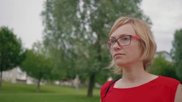 Follow shot of blonde caucasian woman wearing glasses in red dress walking in the city park. Clip re