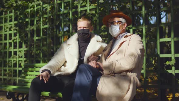 Portrait of Elegant Aged Man and Stylish Young Guy Wearing Safety Mask and Sitting on Bench Outdoors