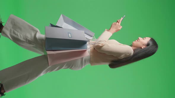 Side View Of Woman With Shopping Bags Using Mobile Phone While Walking In Front Of Green Screen