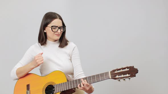 Woman In Beige Outfit Playing Chords On Her Acoustic Nylon Strings Guitar On A Grey Background