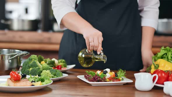 Hands of Female Cook in Apron Pour Olive Oil on Fresh Vegetable Mix