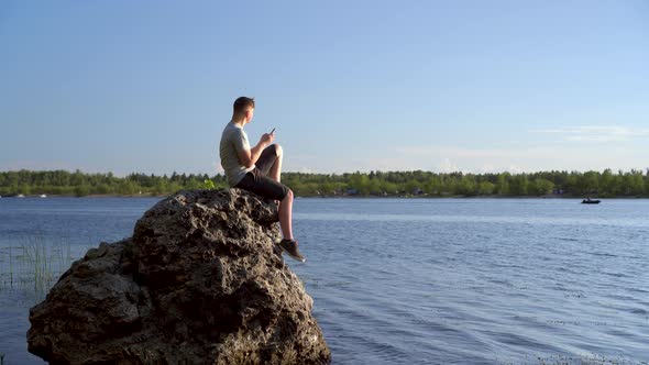 A Young Man Sits on a Stone By the River with a Phone in His Hands. The Man Is Texting on His