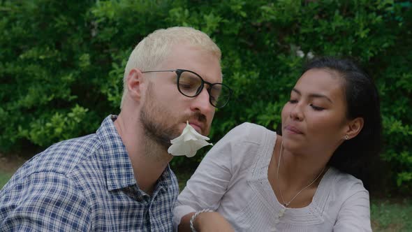 A Young International Couple in Love Sitting on the Grass the Man Holding a Flower in His Teeth and