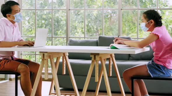 Father and daughter wear protective masks and keep the social distance while working at home.