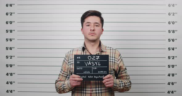 Portrait of Male Criminal in Shirt Raising Head While Holding Sign for Photo in Police Station