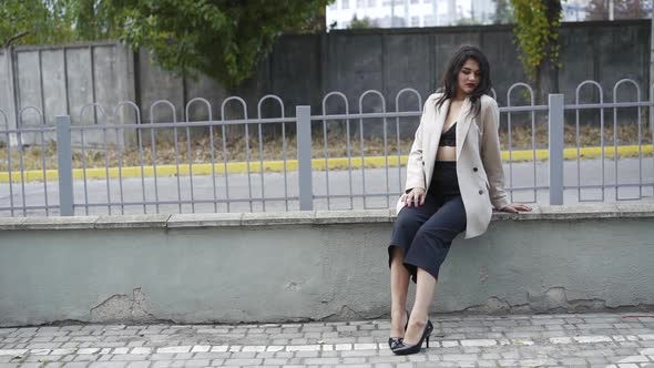 Elegant Brunette in Stylish Clothes Sitting at Fence in Poses with Joy on Face