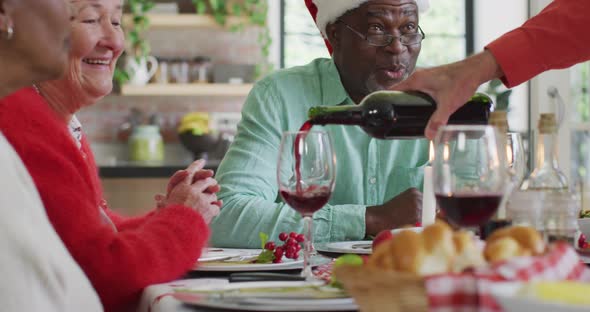 Happy diverse female and male senior friends celebrating meal, pouring vine at christmas time