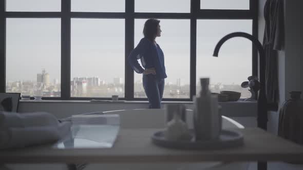 Long Shot of Brunette Caucasian Woman Looking Out the Window and Sitting Down on Windowsill