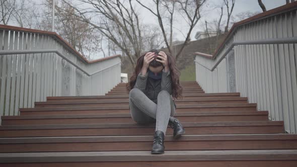 Portrait of Depressed Caucasian Girl Sitting on Stairs in City Park and Crying. Beautiful Brunette