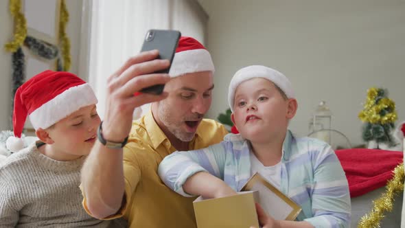 Caucasian father and two sons opening gifts while having a videocall on smartphone during christmas
