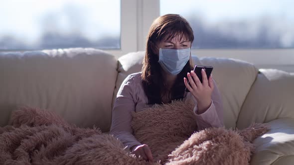 Alarmed Woman in a Mask Sits on the Couch and Calls on the Phone, No Answer