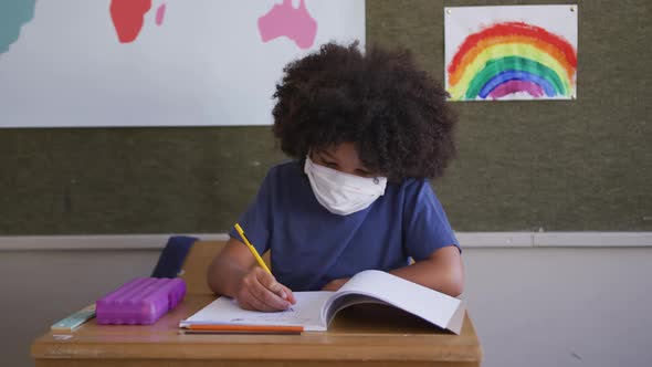 Boy wearing face mask writing while sitting on his desk at school 