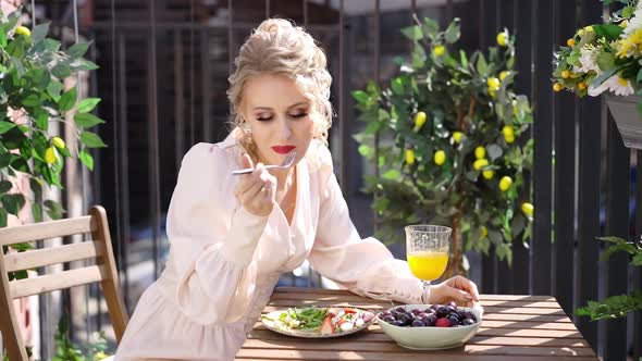 Beautiful Young Woman Eating Salad and Drinking Fresh Juice While Sitting on Balcony on Sunny Day