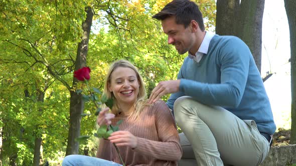 A Boyfriend Comes Up To His Girlfriend Who Sits in a Park, Gives Her a Rose and They Hug