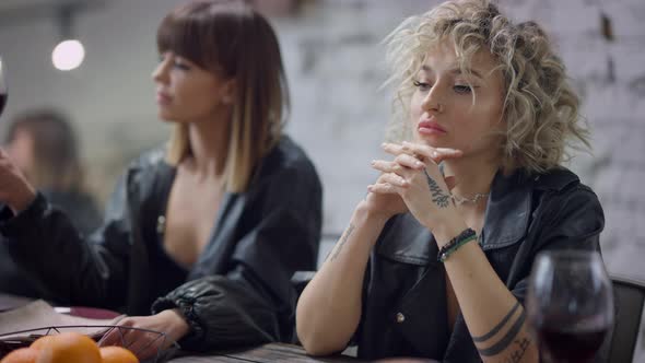 Thoughtful Desperate Lesbian Woman Sitting Indoors with Blurred Partner Drinking Wine at Background