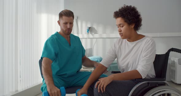 Doctor Helping Patient in Wheelchair with Dumbbells Doing Rehabilitation Exercise in Medical Center