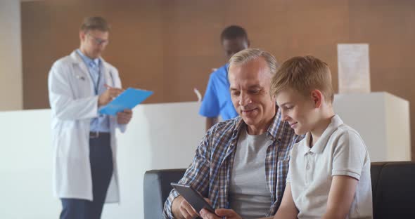 Father and Son Using Digital Tablet Sitting in Hospital Waiting Room