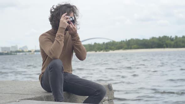 Creative Handsome Man Sitting on River Bank with Retro Camera and Taking Photos. Portrait