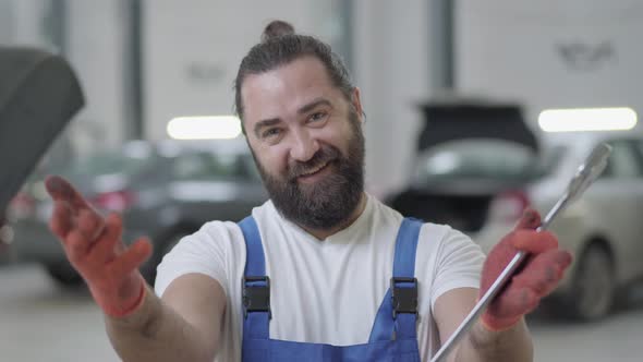 Portrait of Successful Mechanic Makes Inviting Gestures and Smiles Holding Huge Wrench in Hands