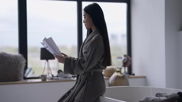 Young Slim Asian Woman Looking Through Business Documents Sitting on Bathtub in Bathrobe