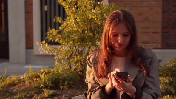 Young attractive business woman sitting outdoor on the bench and using smartphone.