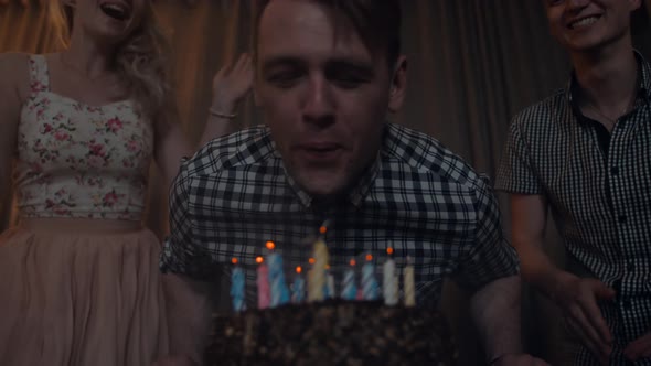 Young Man Blows Out Candles on a Festive Cake with Friends Near.