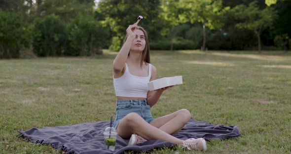 Young Woman in the Park Sitting on a Blue Blanket Picks Up a Susha with Bamboo Sticks and After a