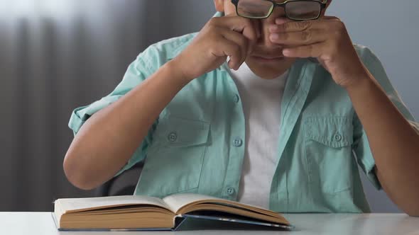 Mixed-Race Male Kid in Glasses Reading Book at Table, Rubbing Eyes, Tiredness