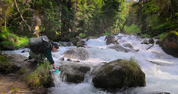 Female Traveler with a Backpack Drinking Water in Nature in the Forest Near a Mountain River