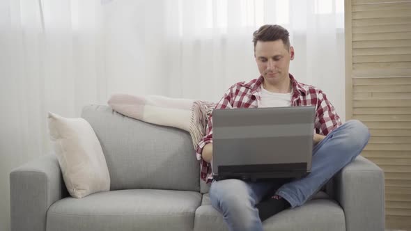 Smiling Brunette Caucasian Man Sitting on Couch and Using Laptop. Portrait of Positive Young Guy