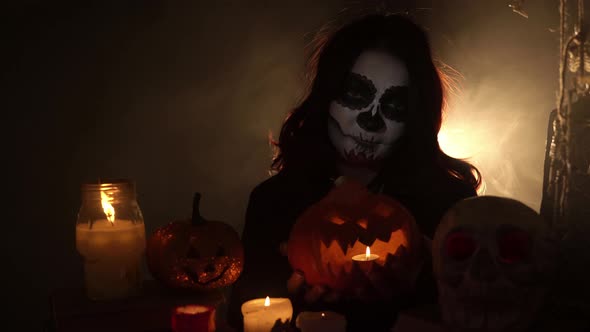 Girl with Skeleton Makeup Sits on a Dark Background and Holds a Lantern Pumpkin