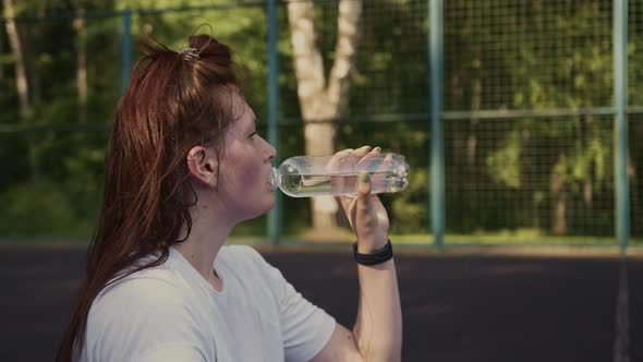 Woman Drinks Water From a Bottle While Sitting on Sports Platform After Training