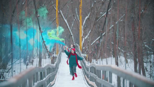 Two Young Happy Women Running on the Snowy Bridge Holding Smoke Bombs