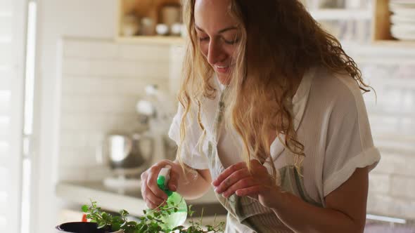 Smiling caucasian woman watering potted plants standing in cottage kitchen