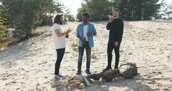 Three Men Stand on the Beach Outside in Front of Them Lies Wood a Bough for a Campfire the Guys Talk