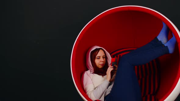 Young Woman Browsing Mobile Phone and Sitting with Her Legs Up in Ballshaped Chair