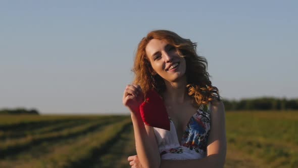 Redhaired Woman in a White Dress Holding a Soft Heart on Background of Field