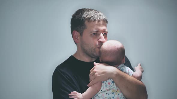 A Father Kisses and Cuddles His Child in His Arms Against a Gray Background