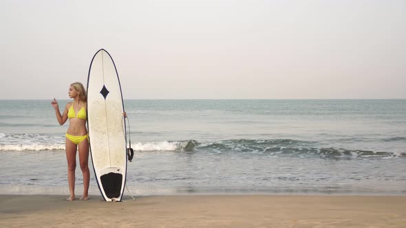 Young Attractive Woman Surfer in a Yellow Bikini with a Surfboard on the Background of the Sea