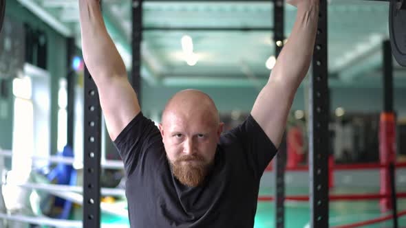 Front View Bald Sportsman with Red Beard Lifting Barbell in Slow Motion Looking at Camera