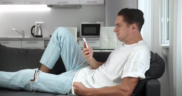 Annoyed Young Man Holding Smartphone Frustrated By Bad Message Resting on Sofa at Home
