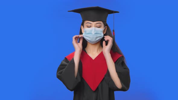Portrait of Female Student in Cap and Gown Graduation Costume Removes Protective Medical Mask