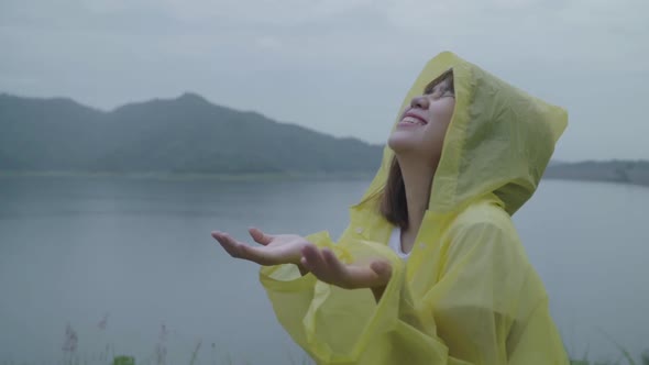 Young Asian woman feeling happy playing rain while wearing raincoat standing near lake.