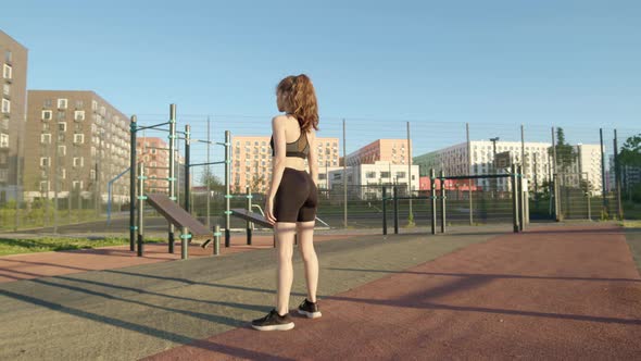 Attractive Athletic Girl in Black Tight Clothes Does Sports Squats on Sports Ground on Street in Her