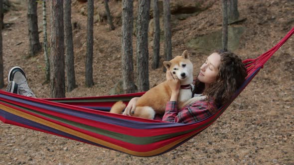 Portrait of Beautiful Young Woman Relaxing in Hammock in Autumn Forest Kissing Shiba Inu Dog