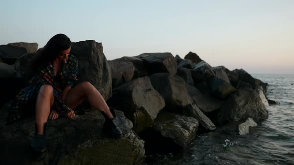 a Brunette in a Swimsuit Shirt and Rough Shoes is Sitting on Large Rocks on the Seashore