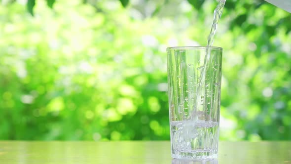 Water From Jug Pouring Into Glass on Wooden Table Outdoors